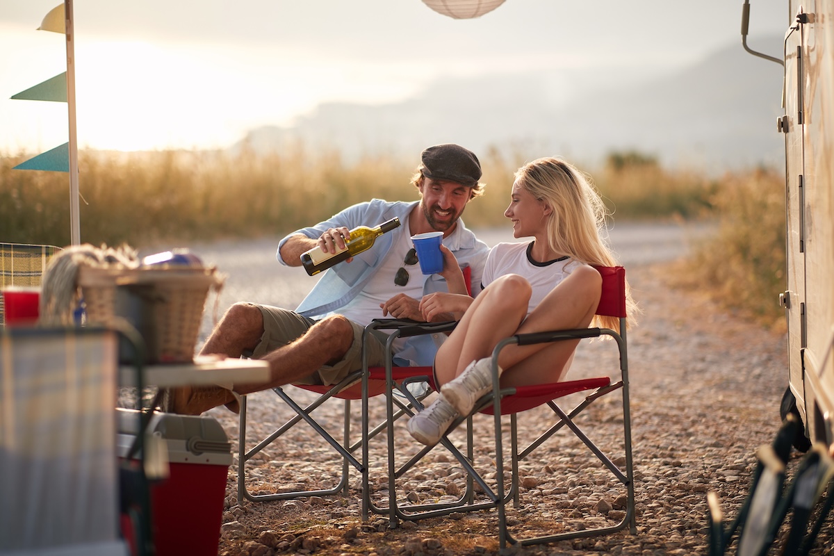 Couple enjoying summertime camping with camper rv in nature, sitting and pouring a glass of wine. Fun, travel, togetherness, love concept.