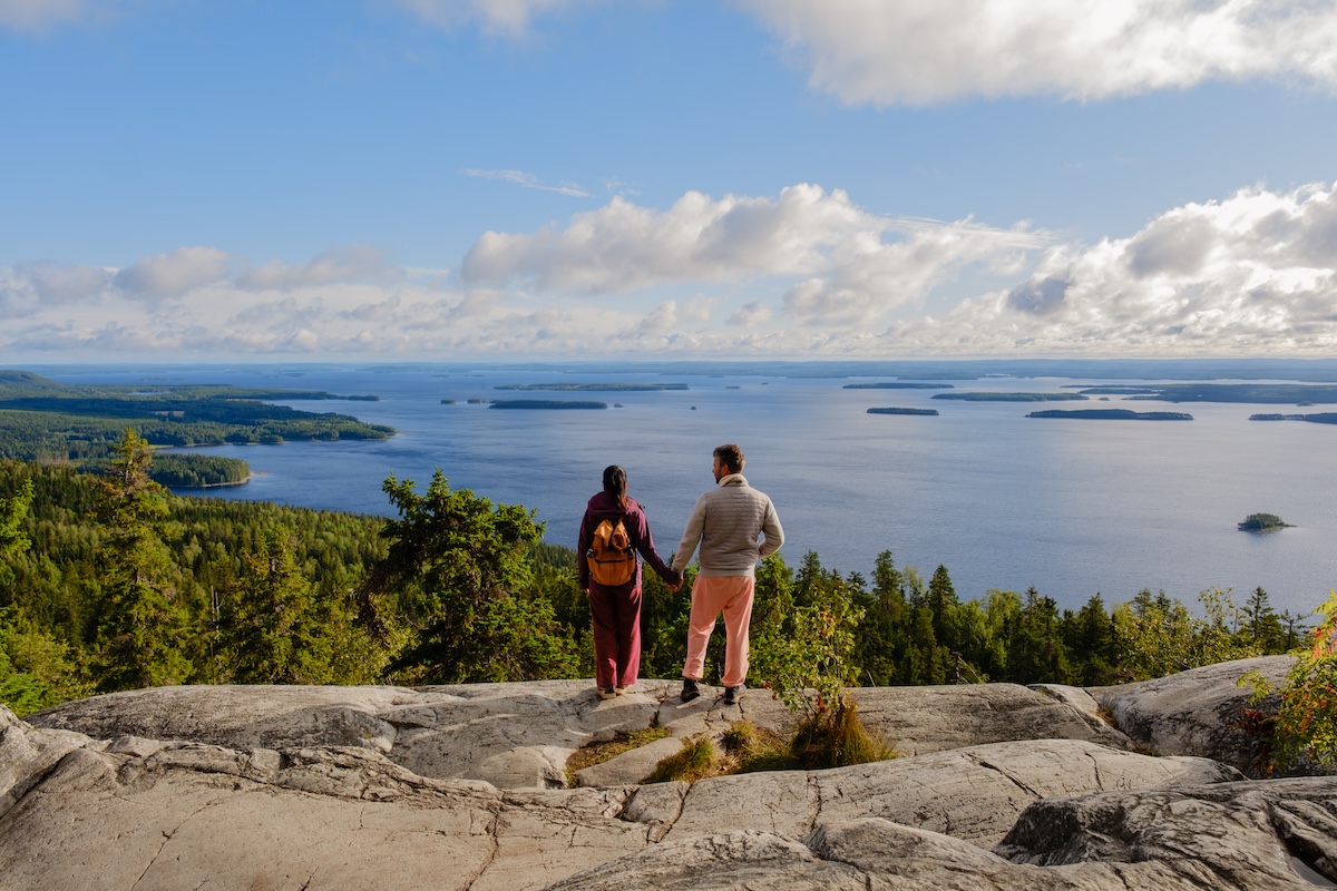 Two hikers stand on a rocky ledge in Koli National Park Finland, admiring the breathtaking view of Lake Pielinen. The clear blue sky and lush greenery enhance the serene atmosphere.